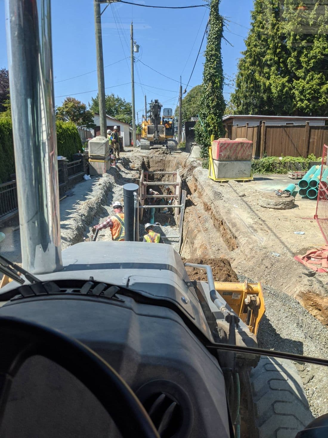 View from a backhoe cab down a shored trench, with a crew laying pipe and an excavator ahead